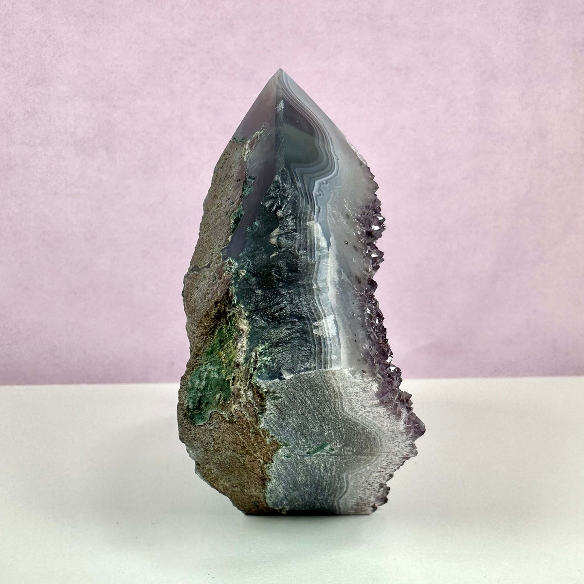 Drusy ametyst spids vist mod en lyserød baggrund. Krystallen har en spids, poleret top med tydelige lag af violette, grå og hvide nuancer samt en glitrende drusy-overflade langs kanten. Den nederste del er ru med naturlige jordtoner og grønne indslag, hvilket fremhæver stenens rå, autentiske udtryk. En harmonisk kombination af skønhed og spirituel energi.