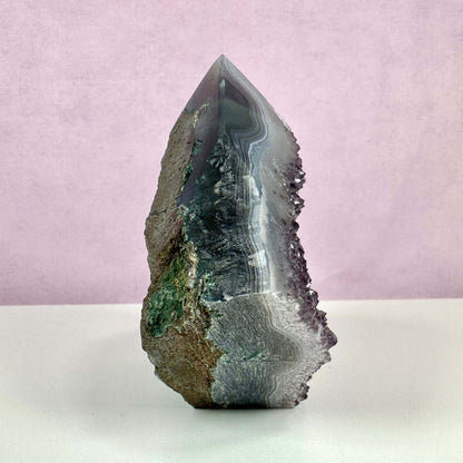 Drusy ametyst spids vist mod en lyserød baggrund. Krystallen har en spids, poleret top med tydelige lag af violette, grå og hvide nuancer samt en glitrende drusy-overflade langs kanten. Den nederste del er ru med naturlige jordtoner og grønne indslag, hvilket fremhæver stenens rå, autentiske udtryk. En harmonisk kombination af skønhed og spirituel energi.