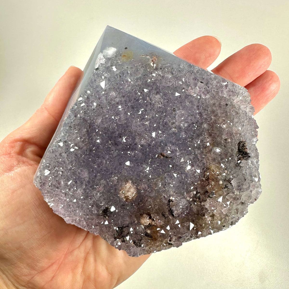 En hånd holder en stor, trekantet drusy ametyst spids med en glitrende overflade af bittesmå krystaller i nuancer af lilla og grå. Krystallen udstråler en naturlig skønhed og intens energi, hvilket gør den ideel til meditation og spirituel praksis. Den ujævne struktur og lysreflekterende krystaller fremhæver stenens unikke æstetiske og helbredende kvaliteter.