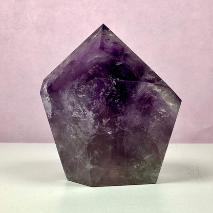 Ametyst spids med dybe violette nuancer, der elegant stråler mod en blød, lyserød baggrund. Den polerede krystal danner en tårnform med naturlige inklusioner, som giver den karakter og dybde. Ametysten symboliserer spirituel indsigt, renhed og beskyttelse samt skaber følelsesmæssig balance. Bruges ofte til meditation, energiarbejde og indretning som en dekorativ og beroligende energikilde.
