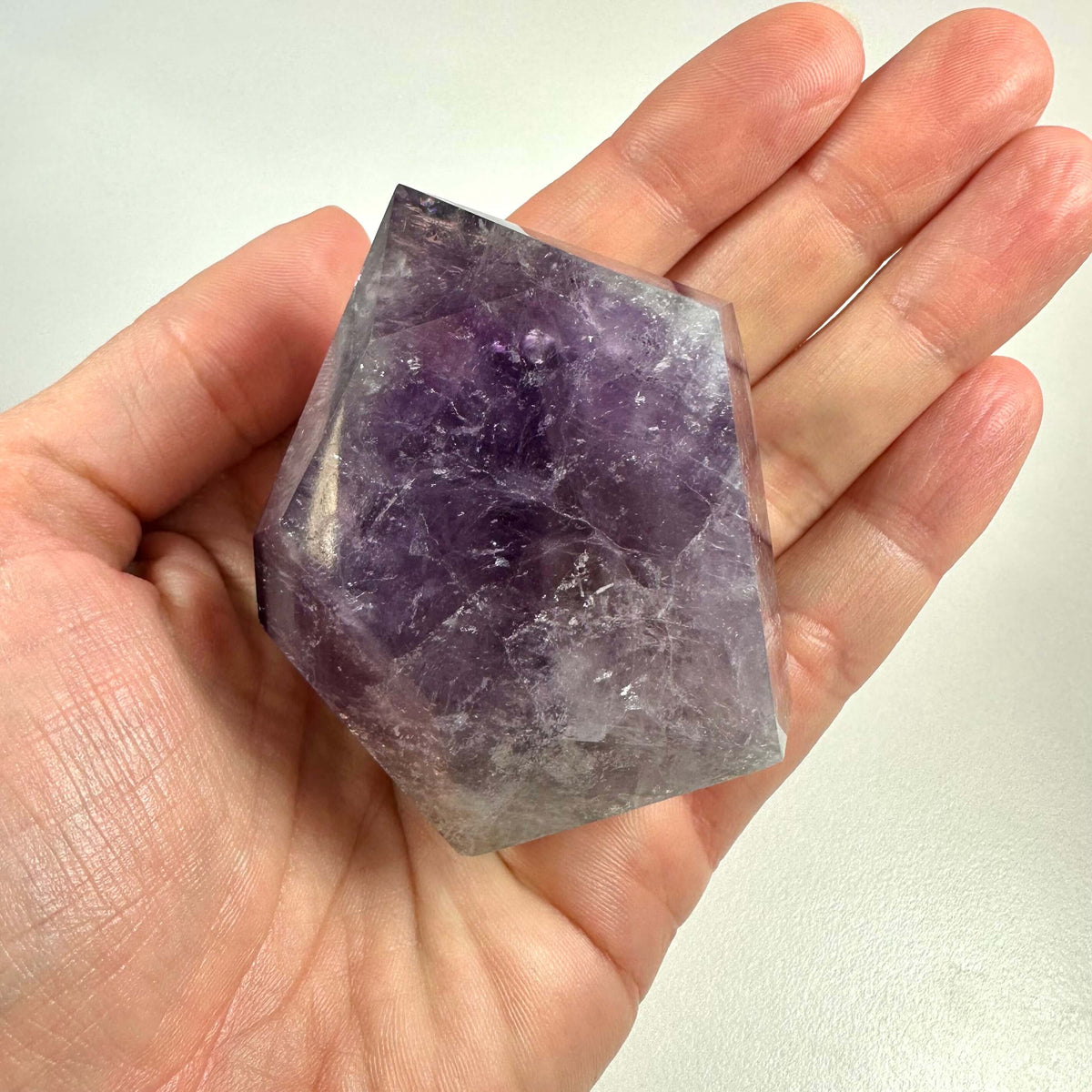 En hånd holder en smukt slebet ametyst spids med tydelige violette nuancer og naturlige inklusioner. Den polerede krystal har en geometrisk form og fremhæver ametystens indre mønstre og farvespil. Ametysten symboliserer åndelig indsigt og beskyttelse og bruges ofte i meditation og energiarbejde. Ideel til både spirituel praksis og som dekorativt element i hjemmet.