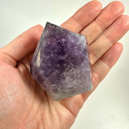 En hånd holder en smukt slebet ametyst spids med tydelige violette nuancer og naturlige inklusioner. Den polerede krystal har en geometrisk form og fremhæver ametystens indre mønstre og farvespil. Ametysten symboliserer åndelig indsigt og beskyttelse og bruges ofte i meditation og energiarbejde. Ideel til både spirituel praksis og som dekorativt element i hjemmet.