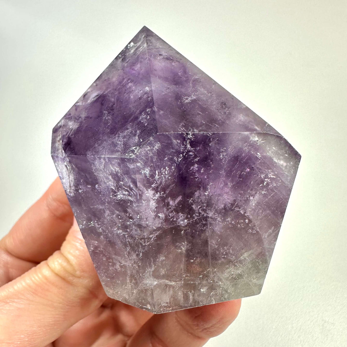 En hånd holder en smukt sleben ametyst spids med klare violette nuancer og karakteristiske naturlige mønstre. Krystallen fremstår poleret i en geometrisk form, der tydeligt fremhæver dens struktur og farvespil. Ametyst spidsen symboliserer åndelig indsigt, indre ro og beskyttelse mod negativ energi. Den er ideel til meditation, energiarbejde og som dekorativt element i hjemmet.