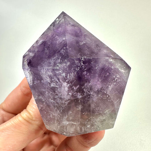 En hånd holder en smukt sleben ametyst spids med klare violette nuancer og karakteristiske naturlige mønstre. Krystallen fremstår poleret i en geometrisk form, der tydeligt fremhæver dens struktur og farvespil. Ametyst spidsen symboliserer åndelig indsigt, indre ro og beskyttelse mod negativ energi. Den er ideel til meditation, energiarbejde og som dekorativt element i hjemmet.