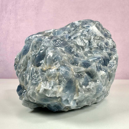Rå blå Calcit nr 1 vises her som en stor, ubehandlet krystal med blå og hvide nuancer, placeret mod en lys baggrund. Stenens overflade er ru og naturlig med tydelige krystalformationer, der fanger lyset smukt. Denne sten er kendt for sin beroligende energi og anvendes ofte til spirituel vækst, følelsesmæssig helbredelse og meditation, især relateret til hals- og tredje øje chakraerne.