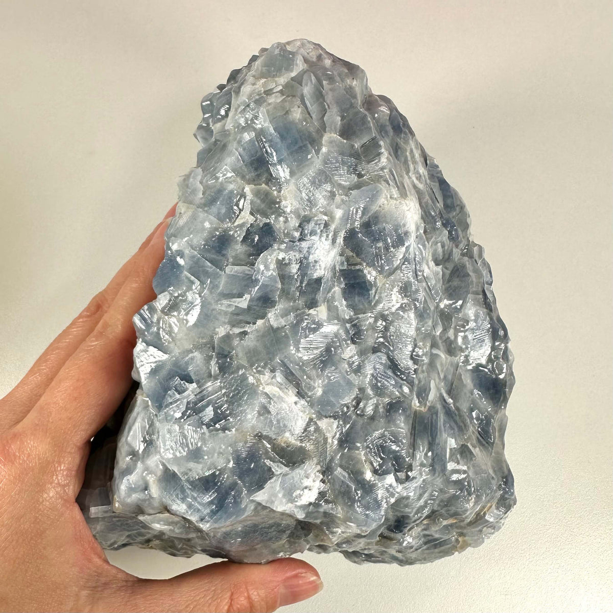 En hånd holder en stor, rå blå Calcit sten med tydelige, skinnende krystaller i nuancer af lys og dyb blå. Stenen har et råt, naturligt udseende og reflekterer lyset på sine mange facetter. Rå blå Calcit nr 1 forbindes med indre ro, følelsesmæssig healing og spirituel vækst, og den arbejder især med halschakraet for at fremme klar kommunikation.