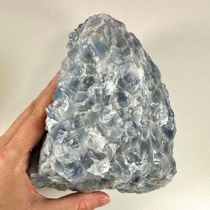 En hånd holder en stor, rå blå Calcit sten med tydelige, skinnende krystaller i nuancer af lys og dyb blå. Stenen har et råt, naturligt udseende og reflekterer lyset på sine mange facetter. Rå blå Calcit nr 1 forbindes med indre ro, følelsesmæssig healing og spirituel vækst, og den arbejder især med halschakraet for at fremme klar kommunikation.