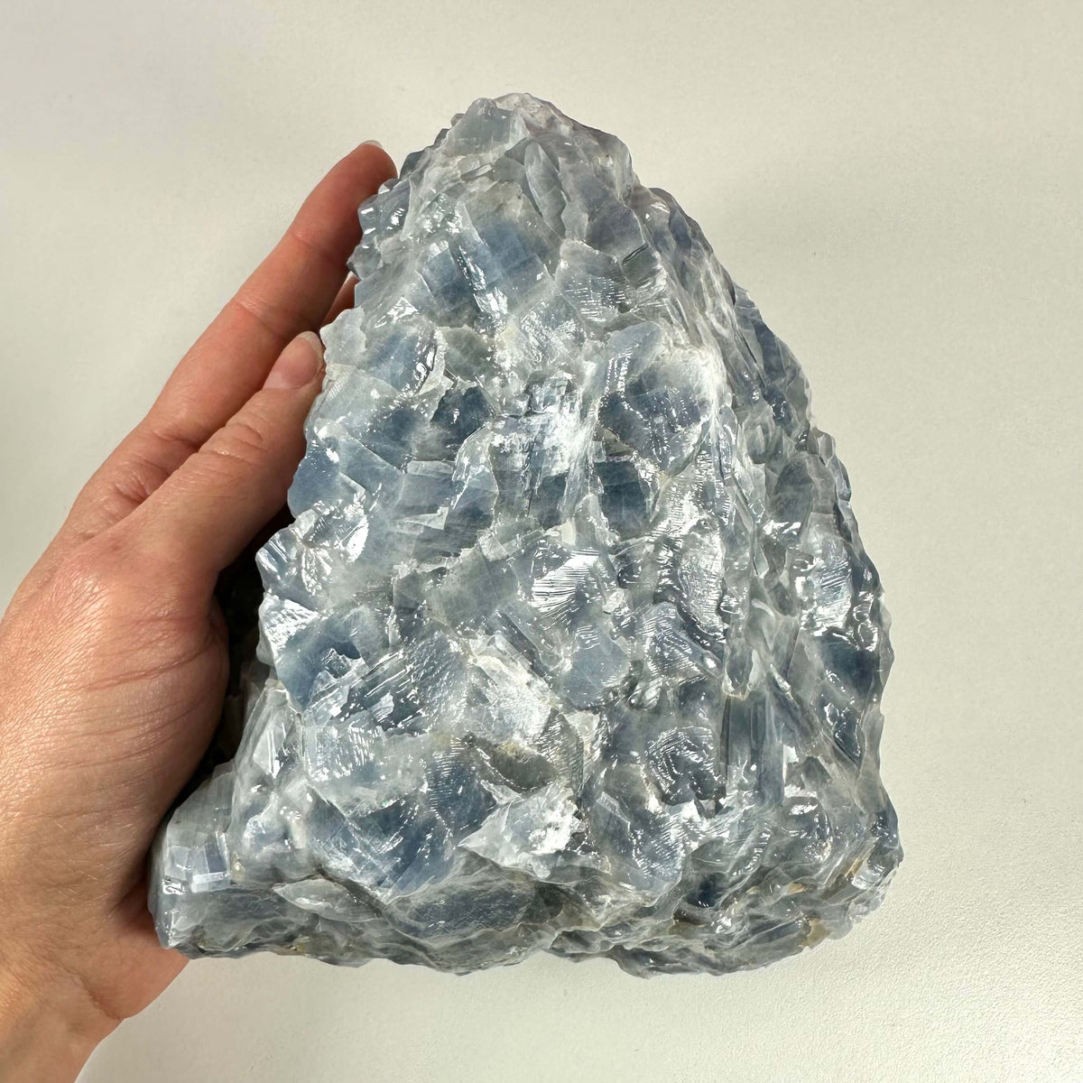 Rå blå Calcit nr 1 vises her som en stor, ujævn krystal i smukke lyseblå og grå nuancer, der holdes i en hånd for at give en fornemmelse af størrelsen. Stenen har en glinsende og naturlig overflade med mange facetter og strukturer. Kendt for sine beroligende energier, bruges blå calcit ofte til spirituel vækst, meditation og styrkelse af halschakraet.