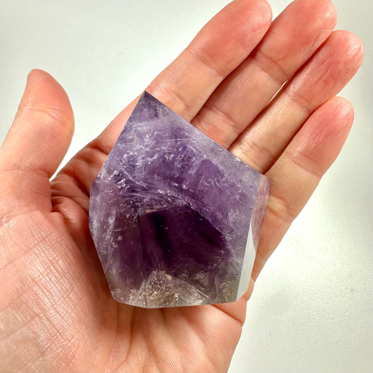 En hånd holder en smuk ametyst spids med tydelige, naturlige lilla nuancer og glitrende krystalstrukturer. Krystallen har en spids form og viser varierende farvedybder fra lys lavendel til mørk violet. Ametyst spidsen symboliserer energi, balance og spirituel indsigt og anvendes ofte til meditation, beskyttelse mod negativ energi samt til at styrke intuition og mental klarhed.