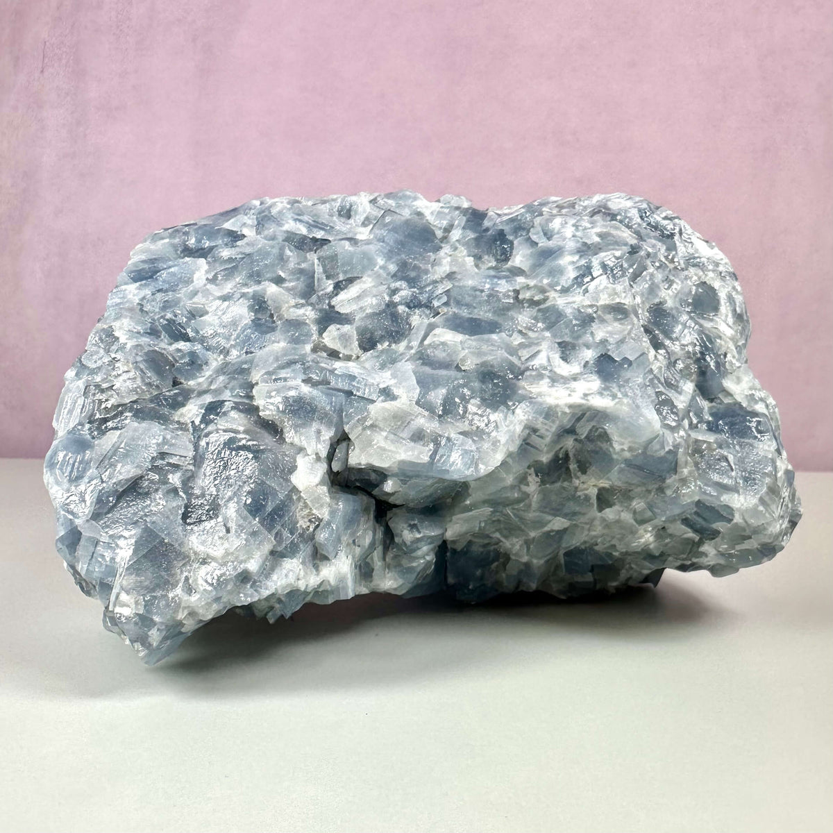 Rå blå Calcit nr 2 er en rå, ubehandlet krystal i nuancer af lys og dyb blå med hvide aftegninger. Den har en porøs, krakeleret overflade og en naturlig, ujævn form. Krystallen er kendt for sine egenskaber, der fremmer ro, følelsesmæssig balance og klar kommunikation. Den vises her mod en lys baggrund og en rosa væg.