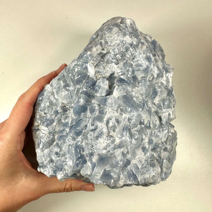 En stor, rå blå calcitkrystal holdes i hånden mod en neutral baggrund. Krystallen har en ujævn, krakeleret overflade med gennemsigtige til mælkehvide og blå nuancer, som fanger lyset smukt. Den naturlige form og farvespil fremhæver stenens beroligende og æstetiske kvaliteter, og illustrerer krystallens evne til at skabe ro, balance og fremme klar kommunikation.