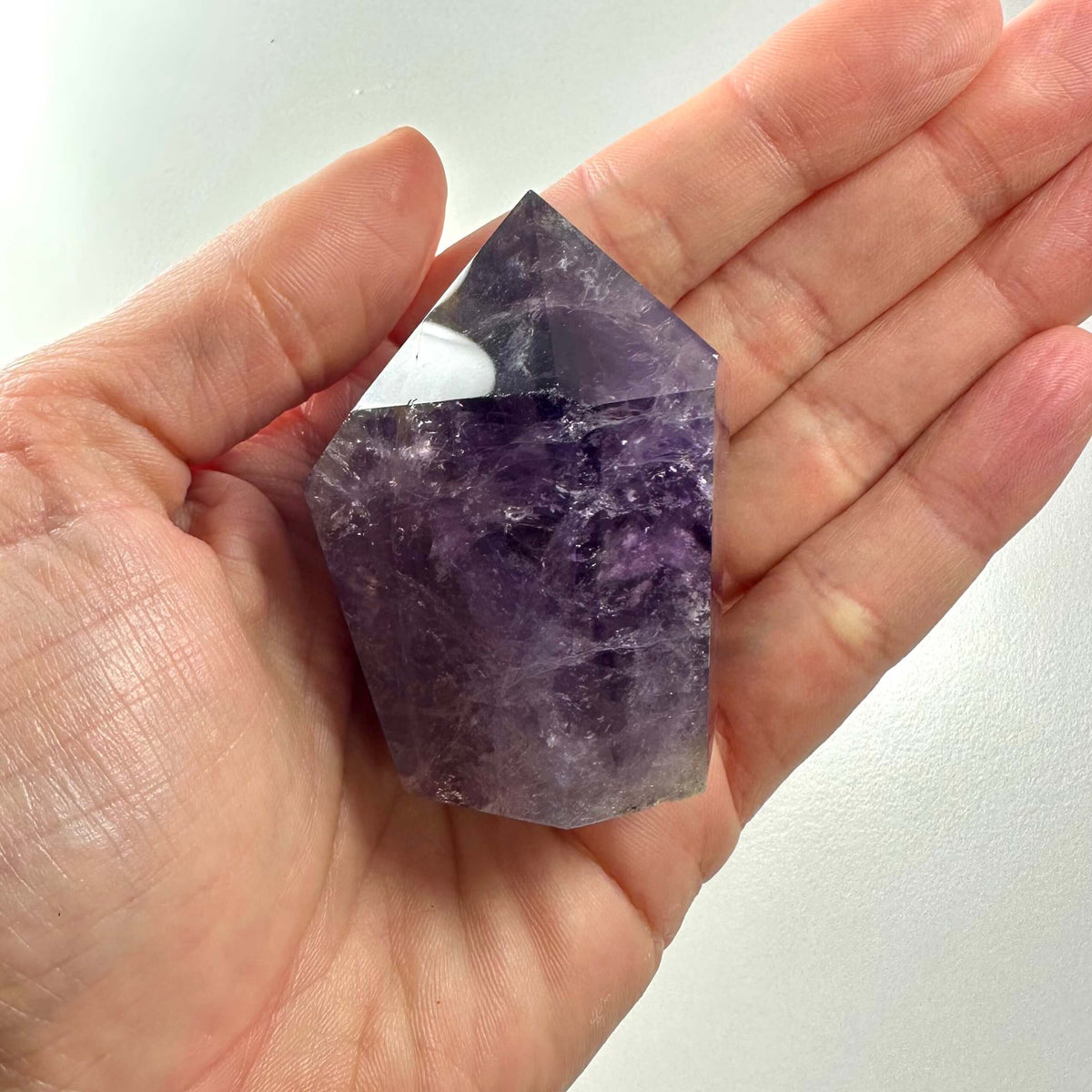 En hånd holder en smukt slebet ametyst spids med dybe lilla nuancer og naturlige indre formationer. Den blankpolerede top og krystallens kraftfulde facon fremhæver dens spirituelle kvaliteter. Ametyst spidsen symboliserer ro, åndelig indsigt og energi, og bruges ofte i meditation og healing. En ideel krystal for dem, der søger balance og beskyttelse mod negativitet.