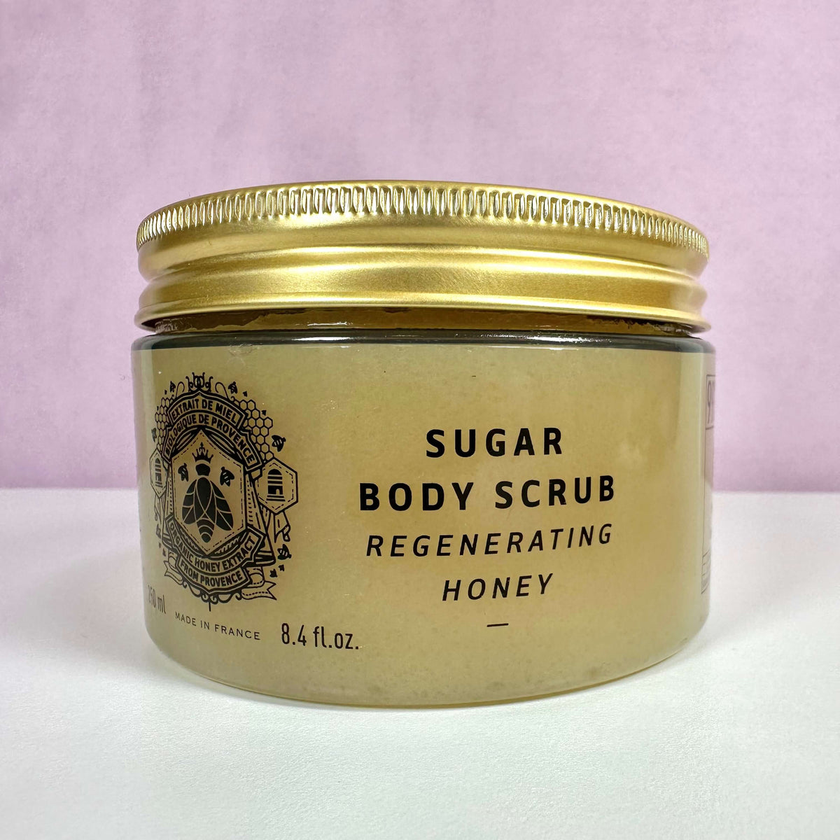 Et close-up billede af Panier Des Sens Sugar Body Scrub – Regenerating Honey. Produktet er præsenteret i en gennemsigtig krukke med gyldent låg og sort tekst på etiketten. Krukken indeholder 8,4 fl. oz. sukkerbaseret bodyscrub med honning, kendt for sine regenererende egenskaber. Baggrunden er enkel med en lys rosa nuance, hvilket fremhæver det elegante og naturlige design.