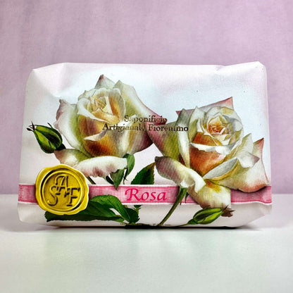 Fiorentino Rosa håndsæbe er elegant indpakket i papir med et smukt design af lyserøde og hvide roser. På forsiden ses teksten "Sapone Artigianale Fiorentino" samt et gult segl med initialerne "SAF". Den romantiske emballage udstråler italiensk luksus og klassisk charme, hvilket understreger sæbens eksklusive kvalitet. En duftende hyldest til naturlig skønhed og forkælelse af hænderne.