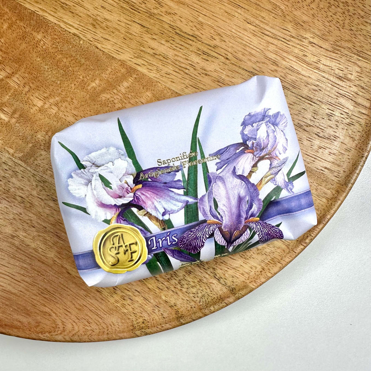 Fiorentino Iris Håndsæbe præsenteres smukt indpakket i papir med illustrationer af delikate irisblomster i lilla og hvide nuancer. Sæbens luksuriøse charme fremhæves af et gyldent segllogo og elegant typografi. Den ligger på et rundt træfad, hvilket understreger produktets naturlige og bæredygtige profil. En harmonisk kombination af skønhed, kvalitet og sanselig velvære.