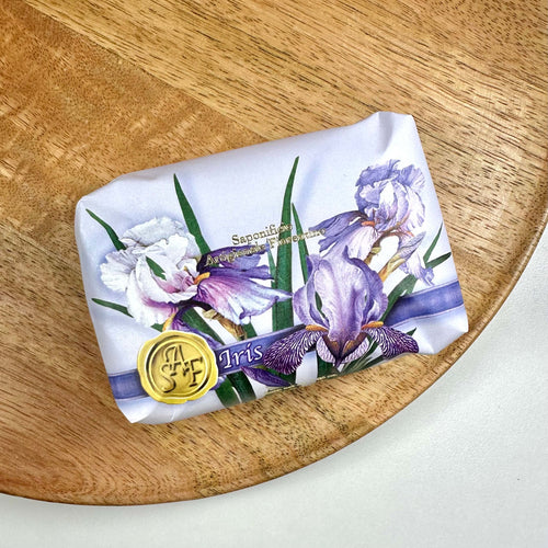 Fiorentino Iris Håndsæbe præsenteres smukt indpakket i papir med illustrationer af delikate irisblomster i lilla og hvide nuancer. Sæbens luksuriøse charme fremhæves af et gyldent segllogo og elegant typografi. Den ligger på et rundt træfad, hvilket understreger produktets naturlige og bæredygtige profil. En harmonisk kombination af skønhed, kvalitet og sanselig velvære.