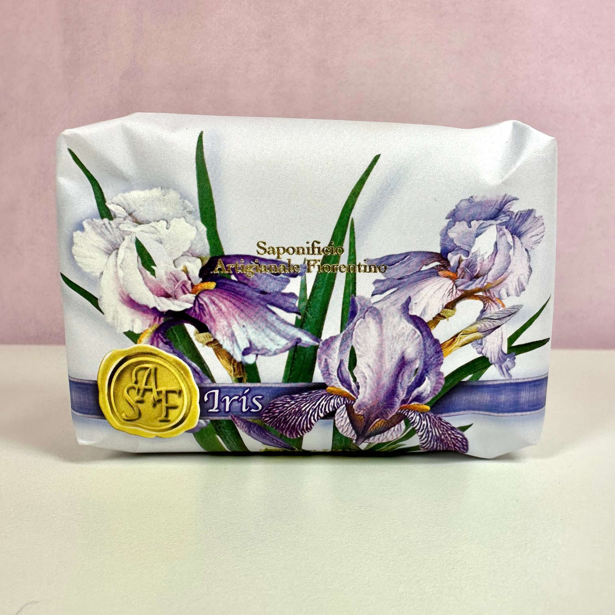 Fiorentino Iris Håndsæbe præsenteres i en elegant indpakning med smukke illustrationer af irisblomster i lilla og hvide nuancer. Produktet signalerer luksus og naturlighed med sin fine grafiske detaljering og subtil guldtekst. Den håndlavede vegetabilske sæbe er ideel til sensitiv hud og tilbyder både fugtgivende pleje og en sanselig duftoplevelse med inspiration fra traditionelt italiensk håndværk.