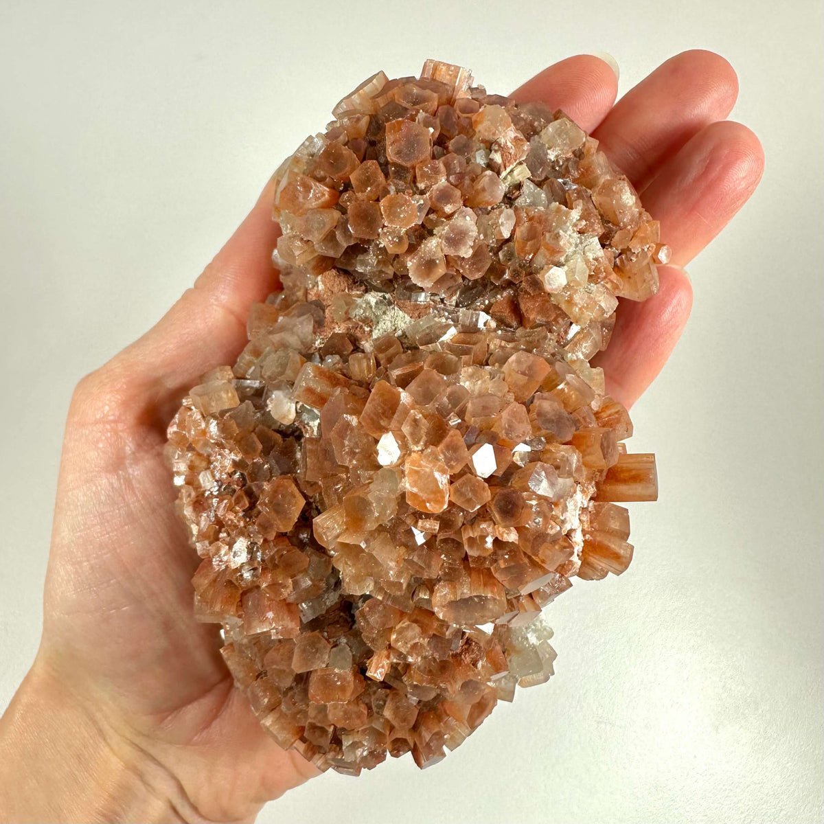 En hånd holder en smuk Aragonit klynge nr. 2 bestående af tæt forgrenede, sekskantede krystalformationer i varme nuancer af brun, gylden og lys orange. Klyngen reflekterer lyset med en naturlig glans og fremstår både rå og detaljerig. Aragonit siges at fremme jordforbindelse, balance og stresslindring, og dens geometriske struktur gør den fascinerende for både samlere og spirituelt interesserede.
