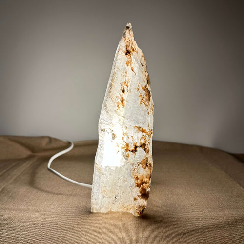 Fé lampe nr 11 er en håndskåret bordlampe lavet af ægte mexicansk selenit, der står på en neutral overflade med baggrund i bløde jordfarver. Lampen udsender et varmt, dæmpbart LED-lys, der fremhæver stenens naturlige lagdelte struktur og gyldne årer. Dens organiske form og beroligende glød skaber en elegant og harmonisk atmosfære i enhver indretning.