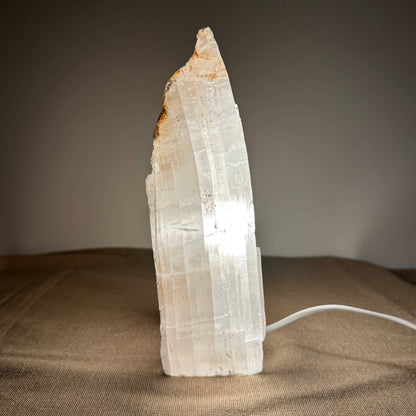 Fé lampe nr. 11 er en håndskåret bordlampe lavet af naturlig selenitkrystal fra Mexico. Lampen har en rå, organisk form og en elegant, hvid overflade, som giver et blødt, dæmpbart lys takket være en integreret LED-pære. Den står på en tekstiloverflade og skaber en rolig og balanceret stemning – perfekt til meditation, afslapning eller hyggelig belysning i hjemmet.