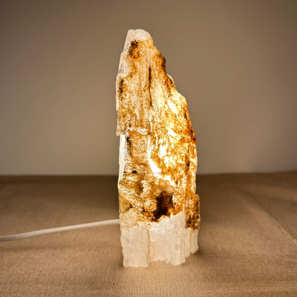 En håndskåret Mexicansk Selenit lampe nr. 48 står oplyst på en neutral overflade med en varm glød, der strømmer indefra. Lampens naturlige, rå struktur og karakteristiske lyse og brune nuancer skaber et organisk og æstetisk udtryk. Denne unikke FÉ lampe kombinerer naturlig skønhed med rensende energi og rolig belysning – perfekt til meditation, afslapning og harmonisk indretning.