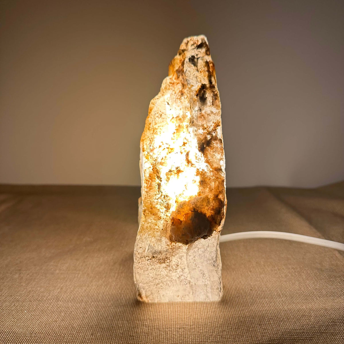 Mexicansk Selenit lampe nr. 48 står oplyst på en neutral overflade og udstråler et varmt, gyldent skær, som fremhæver krystallens naturlige struktur og farvespil. Den håndskårne krystal har organiske former og en rå, ujævn overflade, der skaber et autentisk og æstetisk udtryk. Lampen giver en beroligende atmosfære til rummet og bidrager med rensende energi.