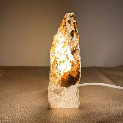 Mexicansk Selenit lampe nr. 48 står oplyst på en neutral overflade og udstråler et varmt, gyldent skær, som fremhæver krystallens naturlige struktur og farvespil. Den håndskårne krystal har organiske former og en rå, ujævn overflade, der skaber et autentisk og æstetisk udtryk. Lampen giver en beroligende atmosfære til rummet og bidrager med rensende energi.
