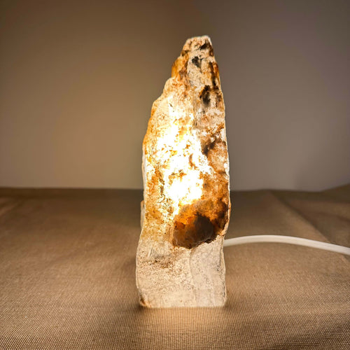 Mexicansk Selenit lampe nr. 48 står oplyst på en neutral overflade og udstråler et varmt, gyldent skær, som fremhæver krystallens naturlige struktur og farvespil. Den håndskårne krystal har organiske former og en rå, ujævn overflade, der skaber et autentisk og æstetisk udtryk. Lampen giver en beroligende atmosfære til rummet og bidrager med rensende energi.