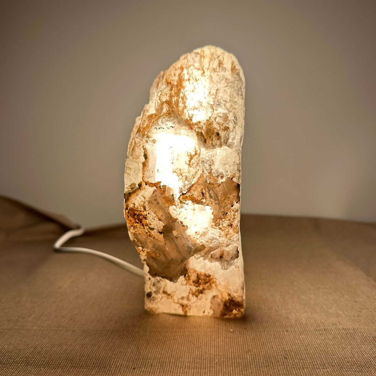 Mexicansk Selenit lampe nr. 10 står oplyst på en neutral baggrund. Den rustikke, håndskårne lampe består af naturlig selenitkrystal med varme jordtoner og et blødt, gyldent lys, som stråler indefra. Lampens unikke struktur og organiske udtryk skaber en rolig og harmonisk stemning, der passer perfekt til både meditation og indretning med sans for æstetik og velvære.