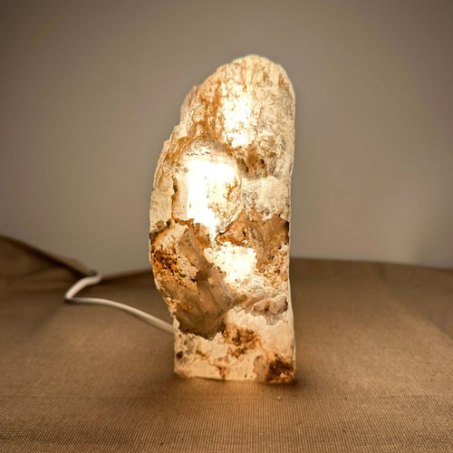 Mexicansk Selenit lampe nr. 10 står oplyst på en neutral baggrund. Den rustikke, håndskårne lampe består af naturlig selenitkrystal med varme jordtoner og et blødt, gyldent lys, som stråler indefra. Lampens unikke struktur og organiske udtryk skaber en rolig og harmonisk stemning, der passer perfekt til både meditation og indretning med sans for æstetik og velvære.