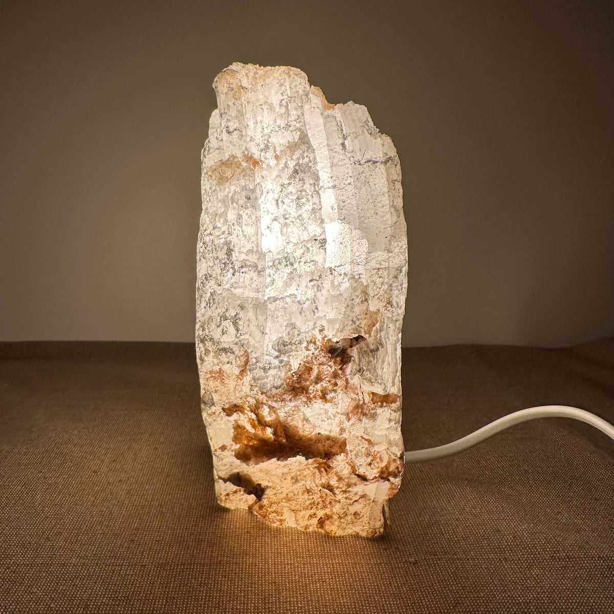 Mexicansk Selenit lampe nr 10 er en håndskåret bordlampe lavet af naturlig selenitkrystal med et råt og organisk udtryk. Lampen spreder et varmt, dæmpbart lys, som skaber en rolig og harmonisk atmosfære. Den står på et neutralt underlag og har en synlig hvid ledning. Den naturlige farve og struktur gør hver lampe unik og ideel til ethvert interiør.