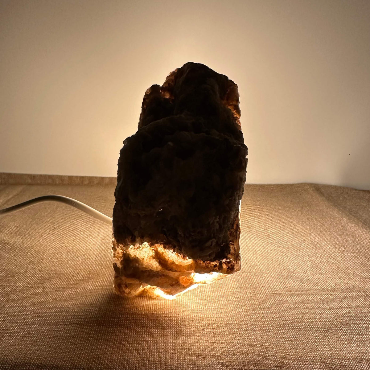 Mexicansk Selenit lampe nr. 30 står oplyst i et dæmpet miljø og afslører sin naturlige, rå tekstur med en varm glød fra bunden. Lampens organiske form og den bløde belysning skaber en beroligende atmosfære. Den er håndskåret af ægte selenitkrystal fra Mexico og kombinerer æstetik med energirensende egenskaber. Perfekt til både meditation og indretning.