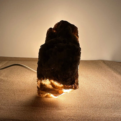 Mexicansk Selenit lampe nr. 30 står oplyst i et dæmpet miljø og afslører sin naturlige, rå tekstur med en varm glød fra bunden. Lampens organiske form og den bløde belysning skaber en beroligende atmosfære. Den er håndskåret af ægte selenitkrystal fra Mexico og kombinerer æstetik med energirensende egenskaber. Perfekt til både meditation og indretning.