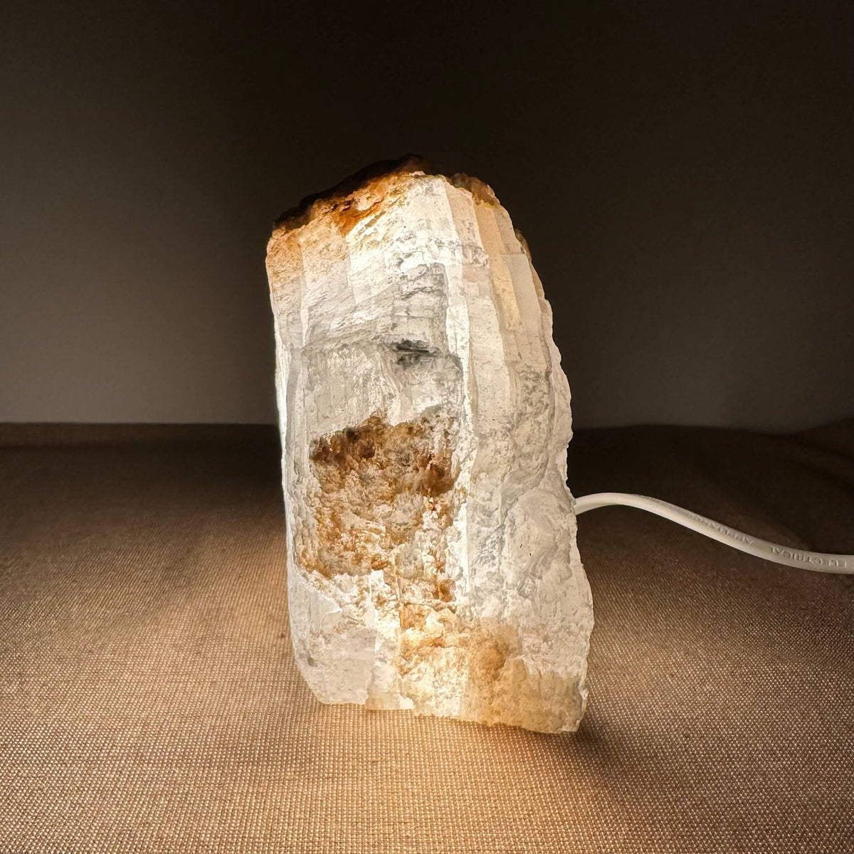 Mexicansk Selenit lampe nr. 30 udstråler et varmt, dæmpet lys fra den håndskårne selenitkrystal med naturlige farver i hvid og gyldenbrun. Lampen står på en neutral overflade og skaber en rolig, stemningsfuld atmosfære. Den unikke struktur og uregelmæssige form af krystallen fremhæver dens naturlige skønhed og gør den til en dekorativ og funktionel lyskilde.