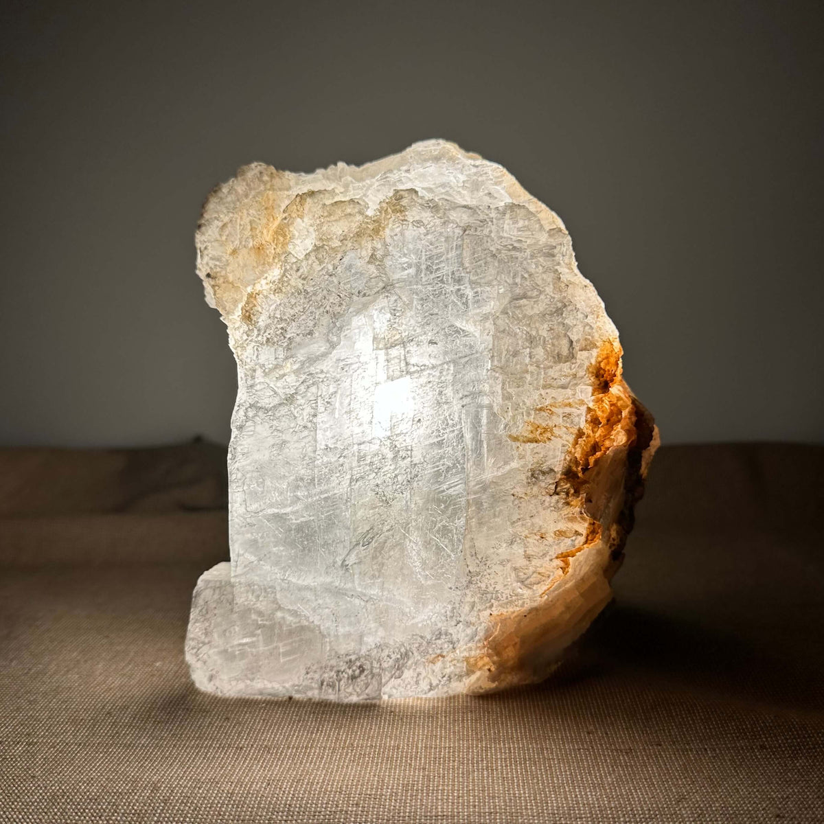 En håndlavet Mexicansk Selenit lampe nr. 7 står oplyst på en brun overflade. Lampen fremviser krystallens naturlige, rå struktur med gennemsigtige og gyldne nuancer, der fanger lyset og skaber en varm, beroligende glød. Lampens organiske, ujævne form vidner om dens unikke oprindelse og æstetik. Perfekt til indretning, meditation og harmonisk belysning i hjemmet.