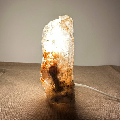 Mexicansk Selenit lampe nr. 7 lyser op i et varmt hvidt skær og fremhæver de naturlige nuancer i den håndskårne krystal. Lampen står på en neutral overflade og udstråler ro og elegance. Den unikke struktur og farvespil i selenitten skaber en organisk og æstetisk tilføjelse til enhver indretning, og understreger lampens naturlige og beroligende egenskaber.