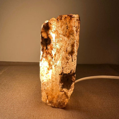 Mexicansk Selenit lampe nr 8 lyser op med en varm glød, som fremhæver dens naturlige, håndskårne krystalstruktur. Lampen står på en neutral overflade og udstråler ro og harmoni. Det unikke design, med jordnære farvevariationer og ru tekstur, skaber en organisk stemning, der passer ind i enhver indretning. Perfekt til meditation, afslapning eller som dekorativ belysning i hjemmet.