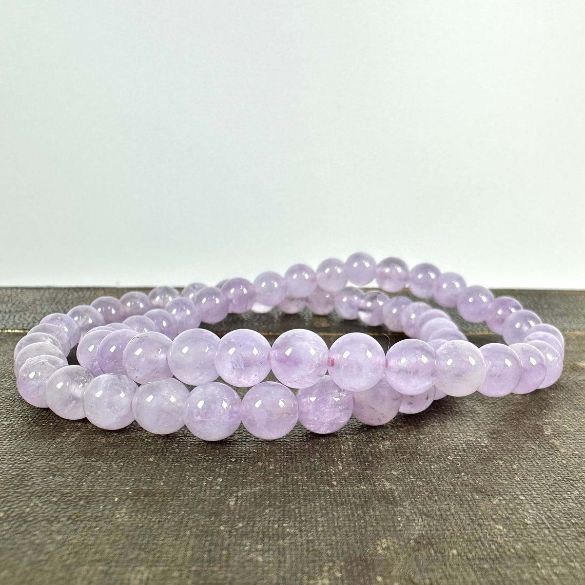 Et elegant Lavendel Ametyst armbånd ligger udstrakt på en mørk overflade med en neutral baggrund. Armbåndet består af glatte, runde perler i en sart lyslilla farve, som minder om lavendelblomster. Den blide nuance og krystallens skær udstråler ro og harmoni. Ideel som smykke og spirituelt redskab, der styrker intuition og fremmer indre balance.