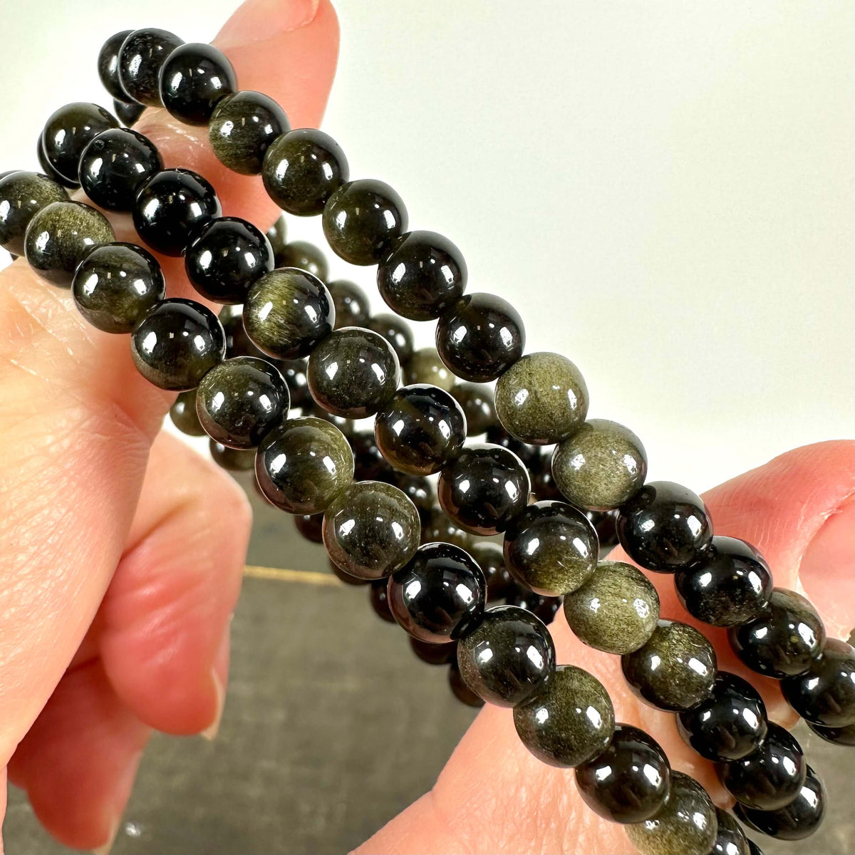 Close-up billede af flere Gylden Obsidian armbånd, holdt mellem to fingre. Armbåndene består af blankpolerede, mørke perler med en gylden glans, som reflekterer lyset smukt. Den gyldne obsidian er kendt for sine helbredende egenskaber, støtte til rodchakraet og evne til at skabe følelsesmæssig balance. Perfekt som smykke og til meditation.