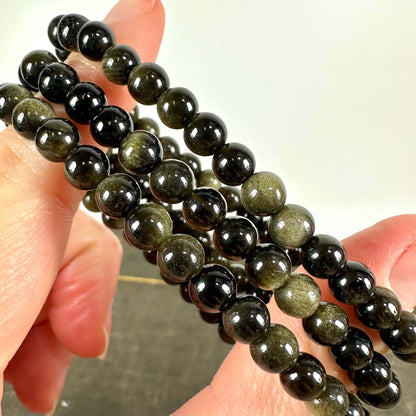 Close-up billede af flere Gylden Obsidian armbånd, holdt mellem to fingre. Armbåndene består af blankpolerede, mørke perler med en gylden glans, som reflekterer lyset smukt. Den gyldne obsidian er kendt for sine helbredende egenskaber, støtte til rodchakraet og evne til at skabe følelsesmæssig balance. Perfekt som smykke og til meditation.