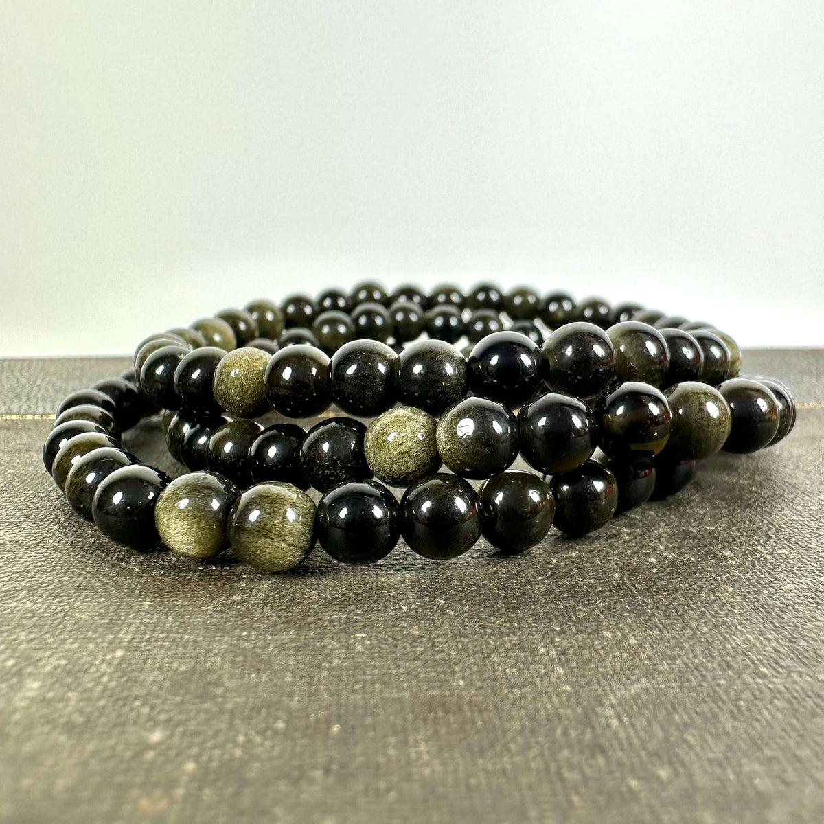 Gylden Obsidian armbånd med smukke, skinnende perler i mørke nuancer og gyldne reflekser, elegant arrangeret i et elastisk bånd. Armbåndet ligger på en rå overflade og fremhæver stenens naturlige glans. Kendt for sine healende egenskaber og stærke forbindelse til rodchakraet, tilbyder dette smykke både energetisk støtte og stilfuld beskyttelse i hverdagen.