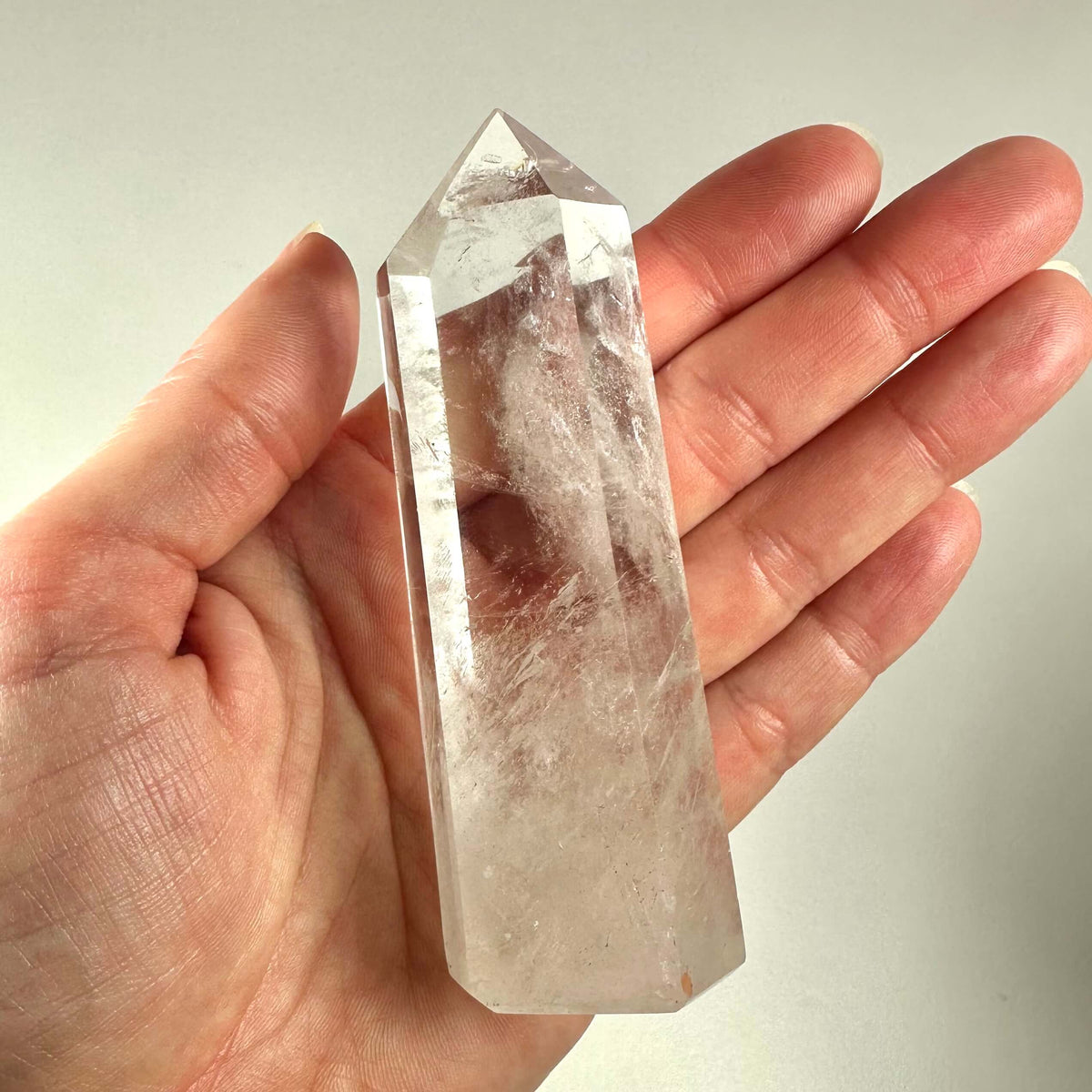En hånd holder et klart bjergkrystal tårn med en glat, poleret overflade og spids ende. Krystallen er gennemsigtig med naturlige indeslutninger og reflekterer lyset smukt. Bjergkrystal tårnet er kendt for sine spirituelle egenskaber og bruges ofte til meditation og energiarbejde. Det symboliserer indre klarhed, healing og balance, og er populært blandt krystalentusiaster og spirituelle udøvere.