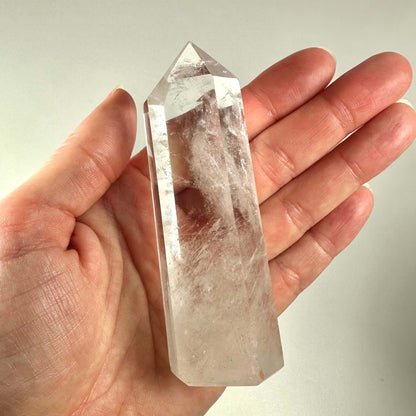 En hånd holder et klart bjergkrystal tårn med en glat, poleret overflade og spids ende. Krystallen er gennemsigtig med naturlige indeslutninger og reflekterer lyset smukt. Bjergkrystal tårnet er kendt for sine spirituelle egenskaber og bruges ofte til meditation og energiarbejde. Det symboliserer indre klarhed, healing og balance, og er populært blandt krystalentusiaster og spirituelle udøvere.
