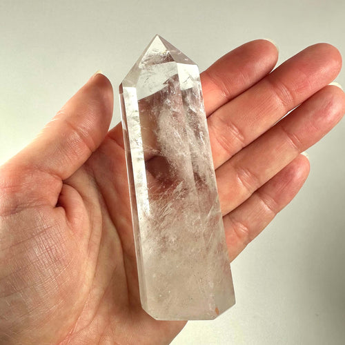En hånd holder et klart bjergkrystal tårn med en glat, poleret overflade og spids ende. Krystallen er gennemsigtig med naturlige indeslutninger og reflekterer lyset smukt. Bjergkrystal tårnet er kendt for sine spirituelle egenskaber og bruges ofte til meditation og energiarbejde. Det symboliserer indre klarhed, healing og balance, og er populært blandt krystalentusiaster og spirituelle udøvere.