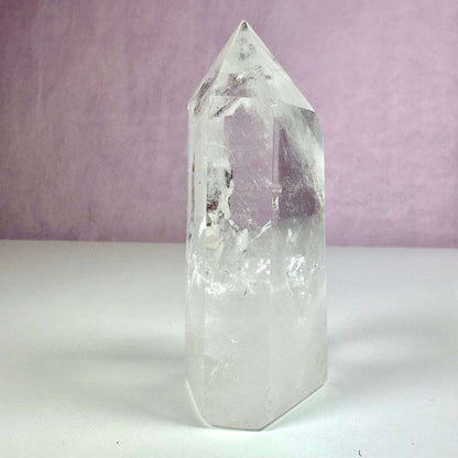 Bjergkrystal tårn af klar kvarts står oprejst mod en lys violet baggrund. Krystallen udstråler gennemsigtighed og indre mønstre, som fanger lyset smukt. Den spidse, slebne top og hexagonale form illustrerer krystallens naturlige struktur. Tårnet symboliserer klarsyn, energibalance og mental klarhed, hvilket gør det ideelt til både dekoration og spirituel praksis i hjemmet.
