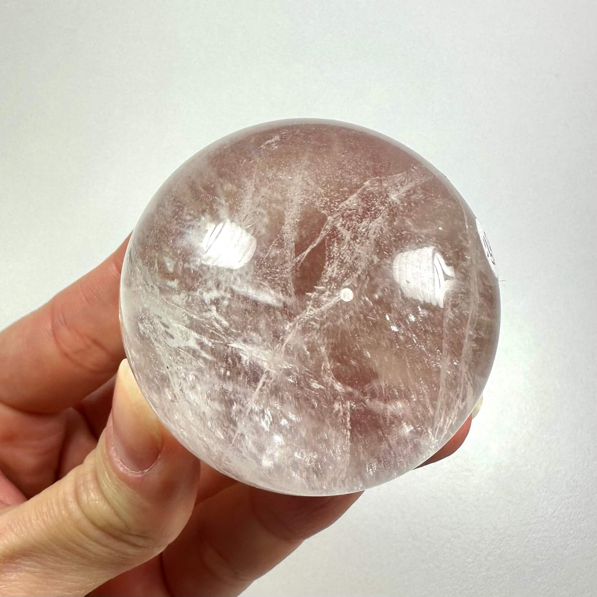 En hånd holder en klar, poleret bjergkrystal kugle op mod en neutral baggrund. Kuglen er gennemsigtig med naturlige inklusioner og indre revner, der reflekterer lyset smukt. Denne smukke krystal symboliserer klarhed, balance og energi-forstærkning. Den anvendes ofte i meditation og healing og anses for at være en kraftfuld energiformidler i spirituelle praksisser.