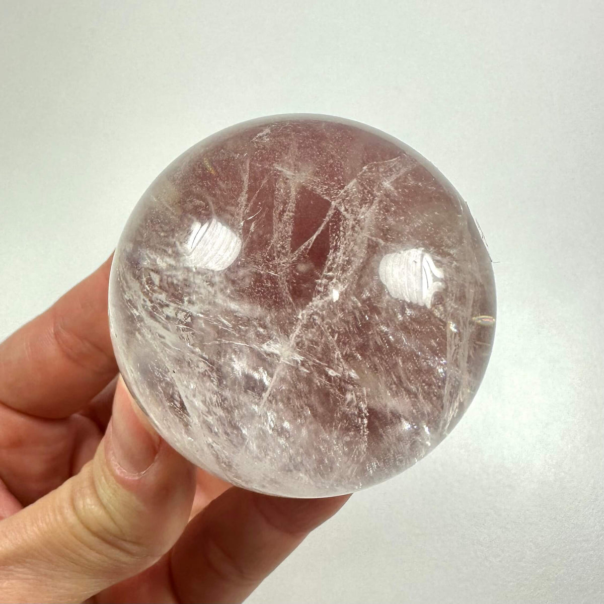 En hånd holder en gennemsigtig bjergkrystal kugle mod en lys baggrund. Kuglen er klar med naturlige indeslutninger og reflekterer lyset smukt. Bjergkrystal symboliserer renhed, klarhed og energi-forstærkning, og anvendes ofte i meditation, healing og spirituelle ritualer. Dens symmetriske form gør den ideel til energiarbejde og afslapning i hjemmet eller under personlige refleksioner.