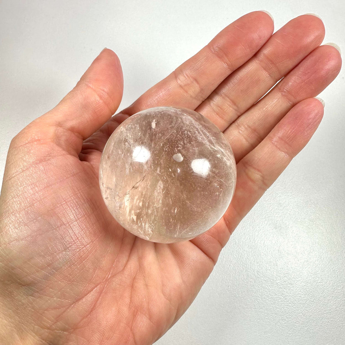 En hånd holder en klar bjergkrystal kugle med lette indre strukturer og reflekser på overfladen. Kuglen er gennemsigtig og rund, og udstråler en følelse af renhed og balance. Bjergkrystal symboliserer klarhed, energi og healing, og bruges ofte i meditation og spirituel praksis som 'mesterhealer'. Den anses for at styrke intentioner og fremme harmoni i både krop og sind.