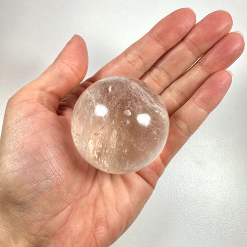 En hånd holder en klar bjergkrystal kugle med lette indre strukturer og reflekser på overfladen. Kuglen er gennemsigtig og rund, og udstråler en følelse af renhed og balance. Bjergkrystal symboliserer klarhed, energi og healing, og bruges ofte i meditation og spirituel praksis som 'mesterhealer'. Den anses for at styrke intentioner og fremme harmoni i både krop og sind.