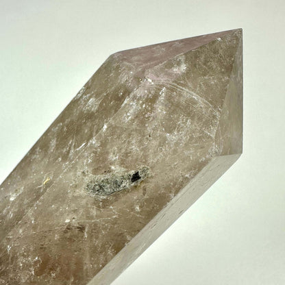 Et nærbillede af et røgkvarts tårn med en poleret, sekskantet form og spids top. Krystallen har en transparent brun til grålig farvetone med naturlige indeslutninger og ujævnheder, som tilføjer karakter. Røgkvarts er kendt for sine jordforbindende og beskyttende egenskaber og bruges ofte i meditation, energihealing og som dekoration i hjemmet eller på arbejdspladsen.